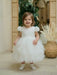 Warm Wendy Off-White Lace Baptism Dress worn by a toddler standing indoors on a light wood floor.