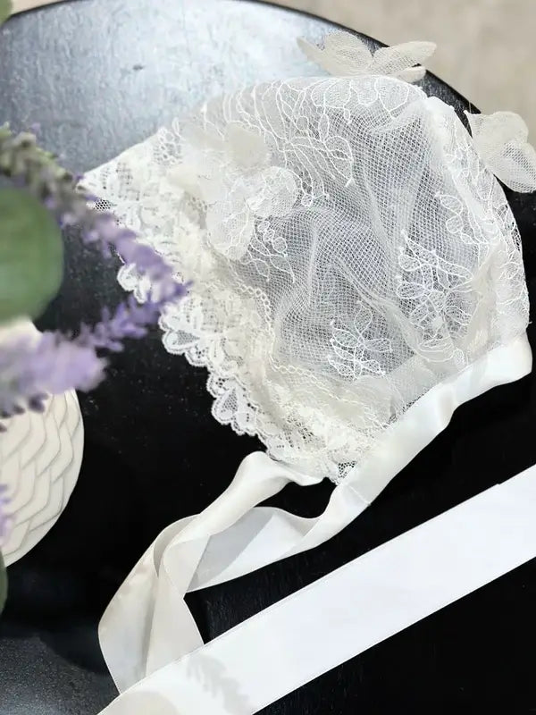 Teter Warm 3D floral detailing baby bonnet with lace and ribbon, displayed on a black surface.