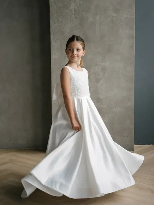 Teter Warm Tara satin bow communion dress shown on a young girl standing indoors against a gray wall.