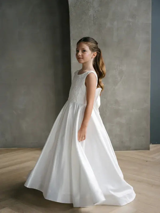 Teter Warm Tara satin bow communion dress worn by a girl standing indoors on a wooden floor.