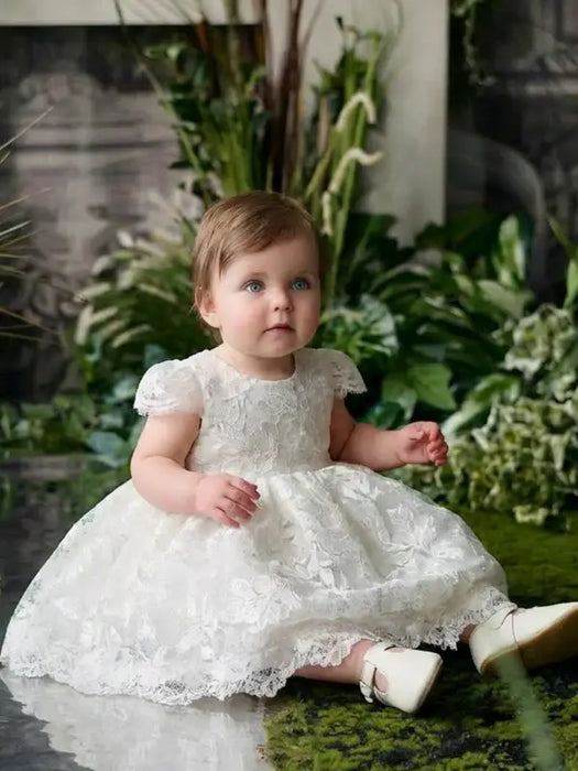 Teter Warm Adele off-white floral lace baptism dress worn by a baby, seated among greenery.