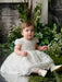 Teter Warm Adele off-white floral lace baptism dress worn by a baby, seated among greenery.