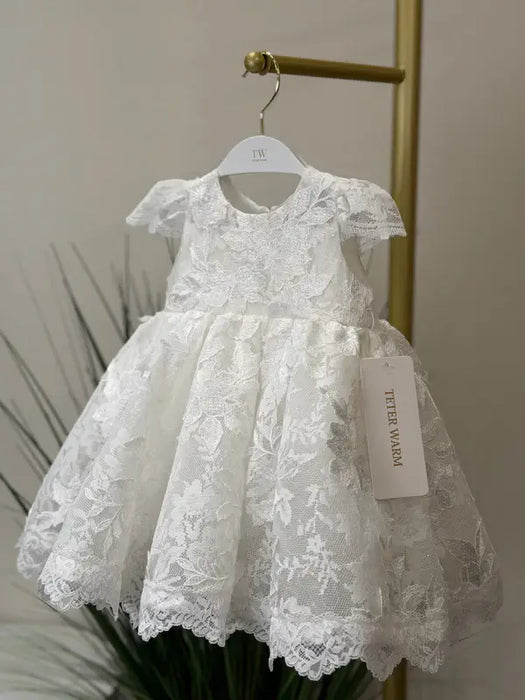 Teter Warm Adele off-white lace baptism dress displayed on a hanger in a softly lit room.