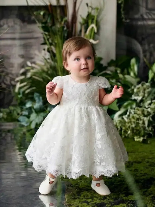 Teter Warm Adele off-white floral lace baptism dress worn by a baby standing by green plants indoors.