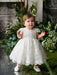 Teter Warm Adele off-white floral lace baptism dress worn by a baby standing by green plants indoors.