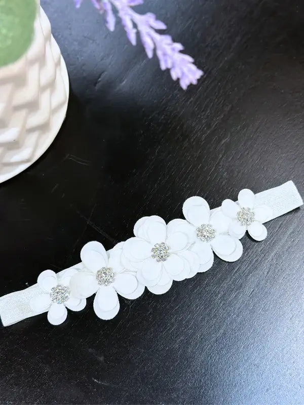 Warm baby girl floral headband with rhinestones on a dark tabletop, featuring white flowers.