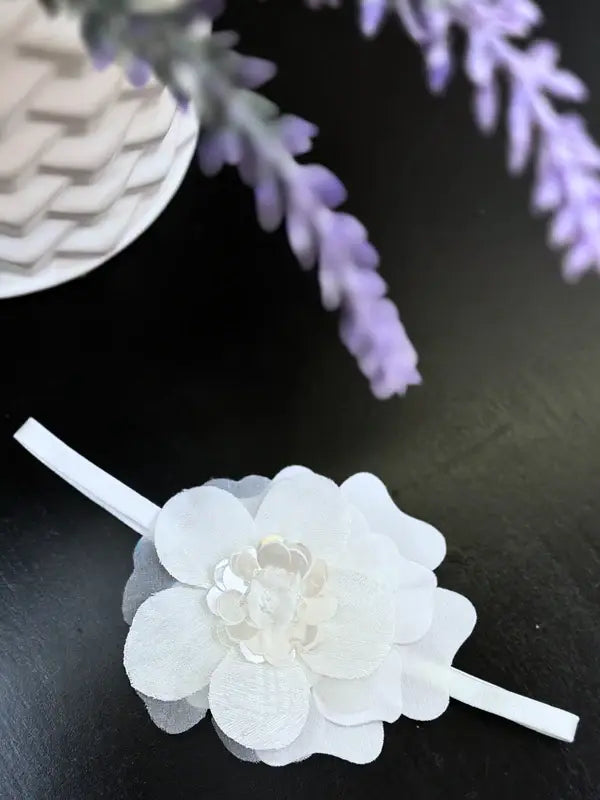 Teter Warm baby girl white flower headband displayed on a dark tabletop beside lavender flowers.
