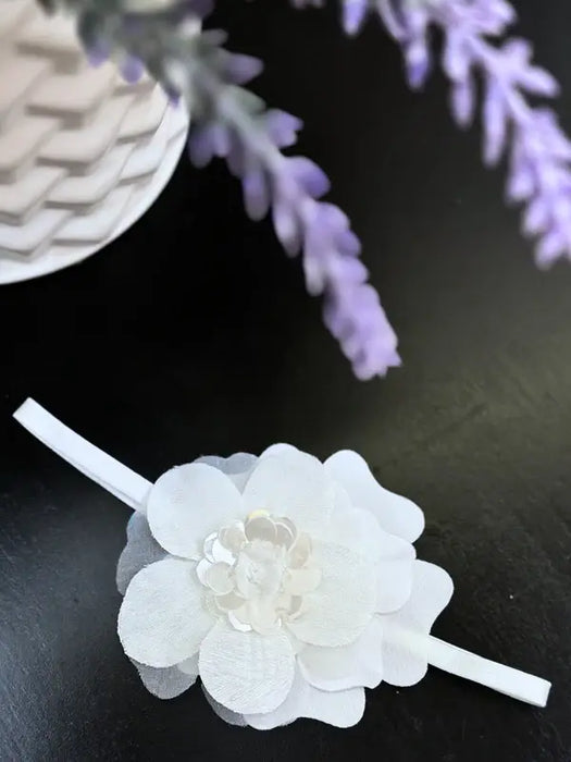 Teter Warm baby girl white flower headband displayed on a dark tabletop beside lavender flowers.