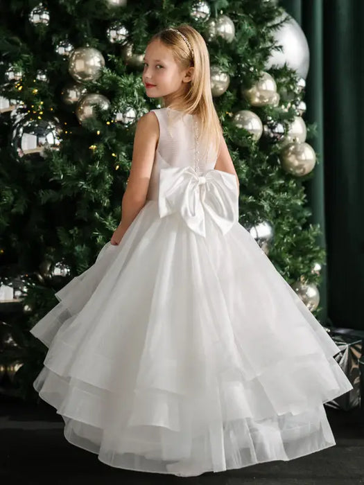 Teter Warm Billie off-white communion dress with bow, shown on a girl standing by a decorated Christmas tree.