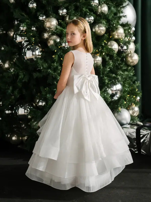 Teter Warm Billie off-white communion dress with bow, worn by a girl standing by a decorated tree.