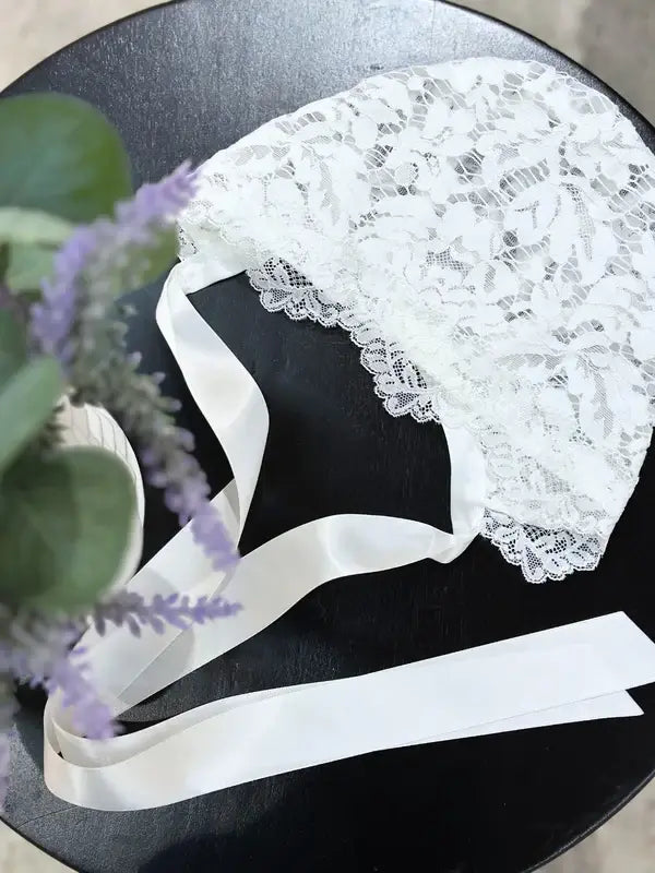 Teter Warm Bold Floral Bay Bonnet white lace baby bonnet with satin ribbons on a dark round table.