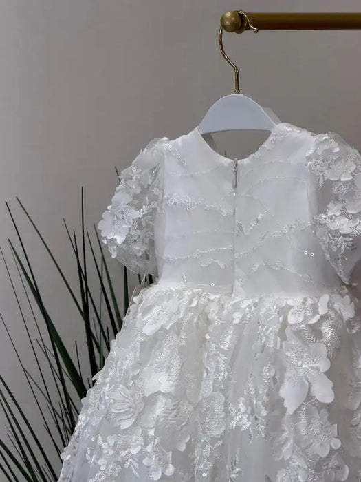 Teter Warm Carlita 3D floral lace baptism dress on a hanger with leafy plants in the background.