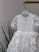 Teter Warm Carlita 3D floral lace baptism dress on a hanger with leafy plants in the background.