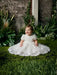 Teter Warm Carlita 3D floral lace baptism dress worn by baby sitting on green grass, surrounded by plants.