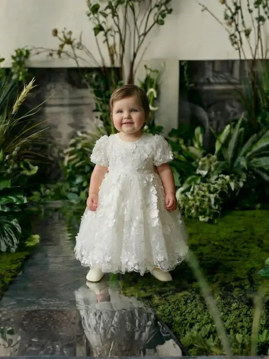 Teter Warm Carlita 3D floral lace baptism dress shown on a toddler standing in a lush garden setting.
