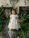Teter Warm Carlita 3D floral lace baptism dress shown on a toddler standing in a lush garden setting.