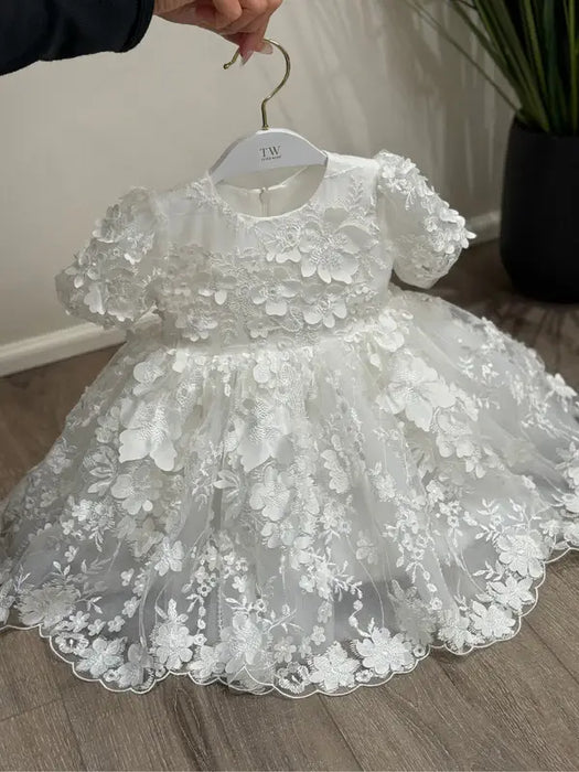 Teter Warm Carlita 3D floral lace baptism dress displayed on a hanger over a wooden floor.