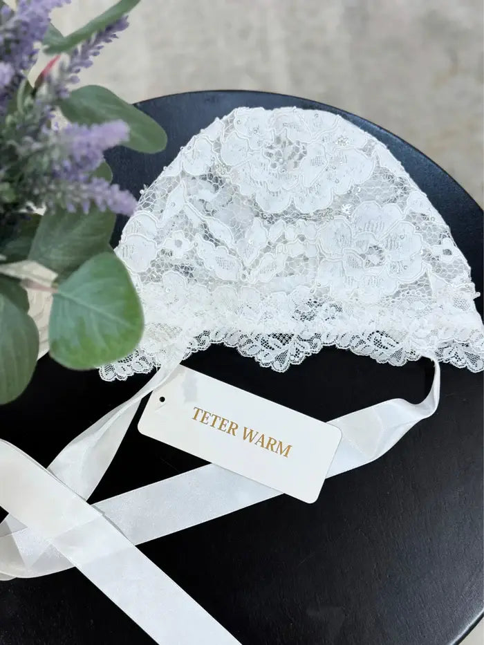 Teter Warm Carol baby bonnet in white lace displayed on a black surface next to a sprig of lavender.