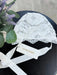Teter Warm Carol baby bonnet in white lace displayed on a black surface next to a sprig of lavender.
