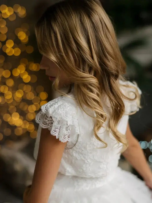 Teter Warm Celeste off-white communion dress with lace sleeves shown on a girl in soft indoor lighting.