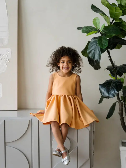Warm Clementine Orange Party Dress shown on a young girl smiling indoors, seated on a modern cabinet.
