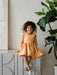 Warm Clementine Orange Party Dress shown on a young girl smiling indoors, seated on a modern cabinet.