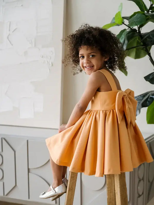 Teter Warm Clementine Orange Party Dress on a smiling child sitting on a wooden stool indoors.