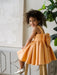 Teter Warm Clementine Orange Party Dress on a smiling child sitting on a wooden stool indoors.