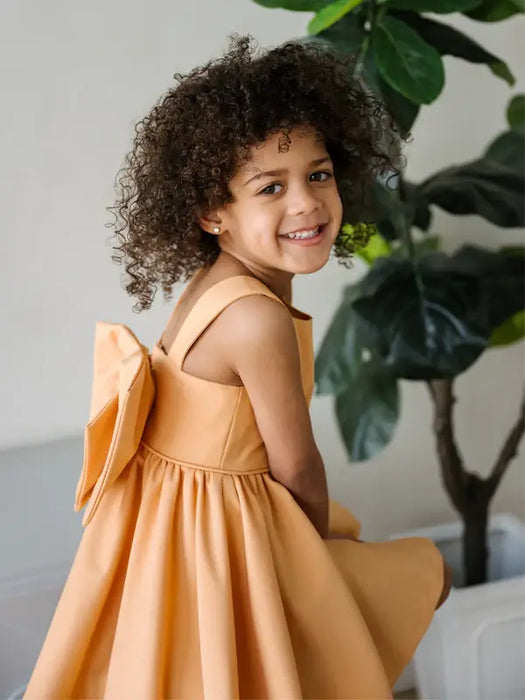 Teter Warm Clementine orange party dress worn by a smiling girl sitting indoors near a plant.