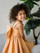 Teter Warm Clementine orange party dress worn by a smiling girl sitting indoors near a plant.