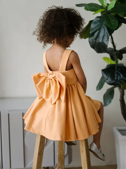 Teter Warm Clementine Orange Party Dress with large bow, shown on a child sitting on a stool indoors.