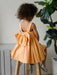 Teter Warm Clementine Orange Party Dress with large bow, shown on a child sitting on a stool indoors.