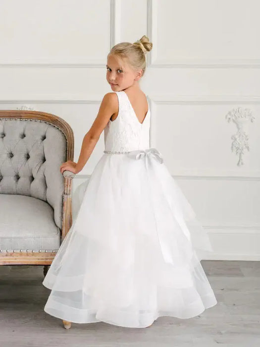 Teter Warm Cora off-white communion dress on a young girl posing beside an elegant chair indoors.