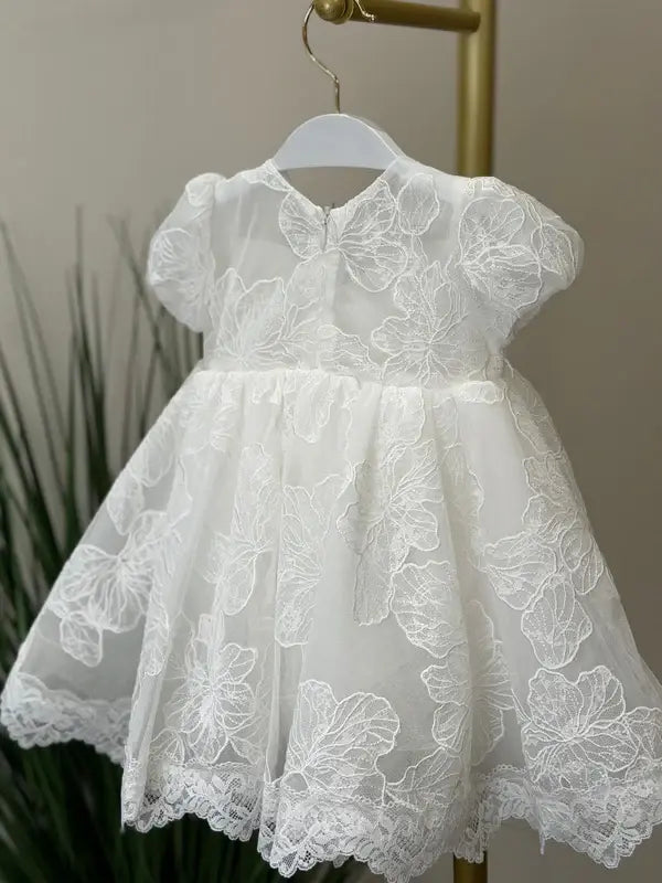 Teter Warm Dalia Floral Off-White Baptism Dress with lace details displayed on a hanger indoors.