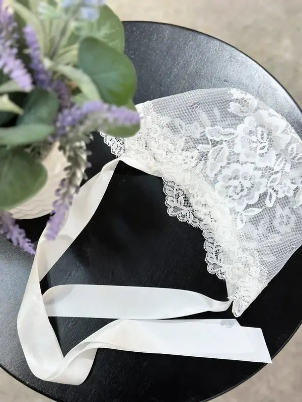 Teter Warm Delicate Baby Bonnet made of white lace with ribbon ties on a black table beside a plant.