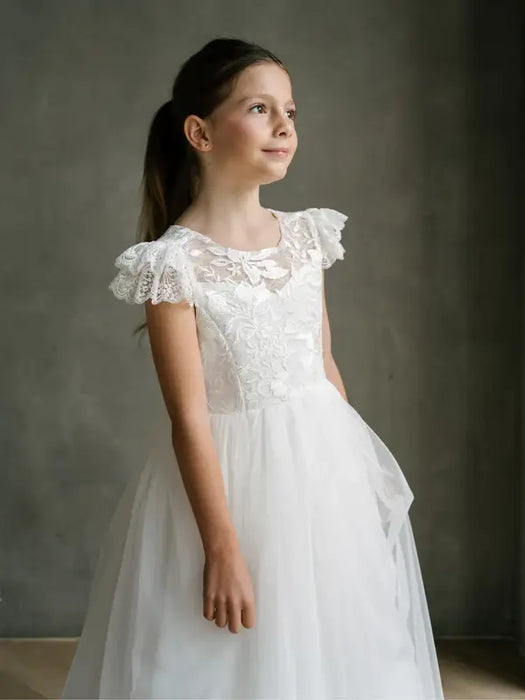 Emma Lace Communion Dress shown on a young girl standing indoors against a neutral background.
