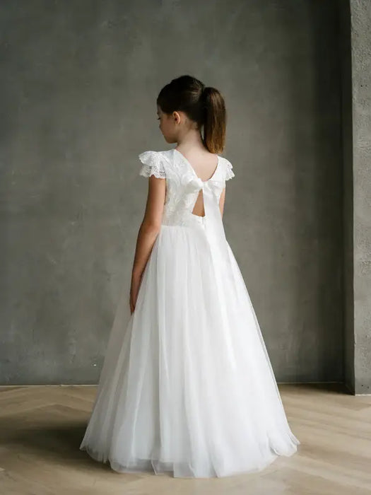 Emma Lace Communion Dress shown on a girl standing indoors, back view highlighting lace detail and bow.