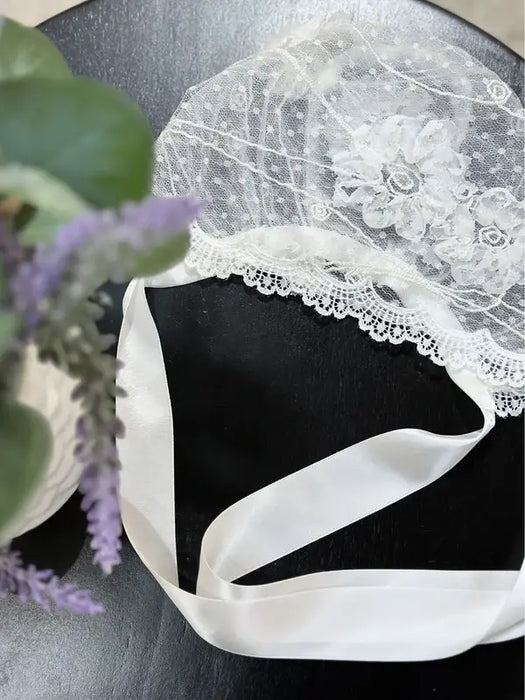 Teter Warm floral lace baby bonnet with satin ties displayed on a dark tabletop.