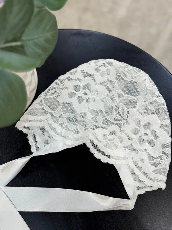 Teter Warm floral lace baby bonnet with satin ties displayed on a dark table.
