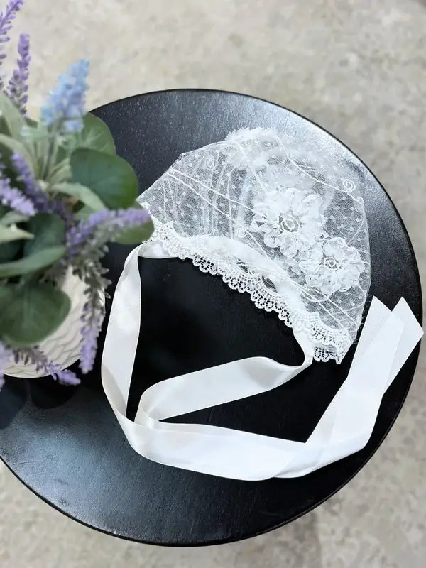 Teter Warm floral lace baby bonnet displayed on a black round table next to a vase of flowers.