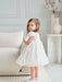 Teter Warm Floral Off-White Lace Baptism Dress worn by a young girl standing near a sofa indoors.