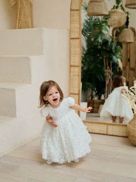 Teter Warm Floral Off-White Lace Baptism Dress shown on a smiling toddler indoors by a mirror.