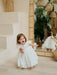 Teter Warm Floral Off-White Lace Baptism Dress shown on a smiling toddler indoors by a mirror.