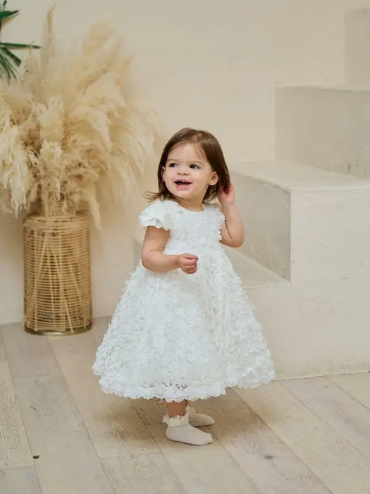 Warm floral off-white lace baptism dress on a toddler standing indoors near stairs.