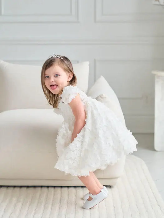 Teter Warm floral off-white lace baptism dress on a smiling toddler standing by a cream sofa.