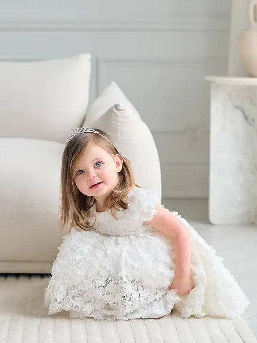 Teter Warm off-white floral lace baptism dress worn by a young girl sitting indoors on a cream rug.