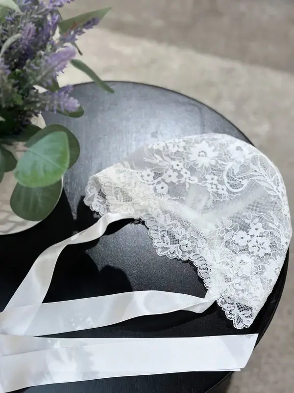 Teter Warm floral soft lace bonnet with satin ribbons displayed on a black table beside a vase of flowers.