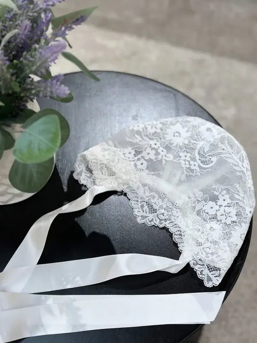 Teter Warm floral soft lace bonnet with satin ribbons displayed on a black table beside a vase of flowers.