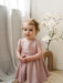 Harper Pink Baby Party Dress shown on a toddler standing indoors near a vase with flowers.