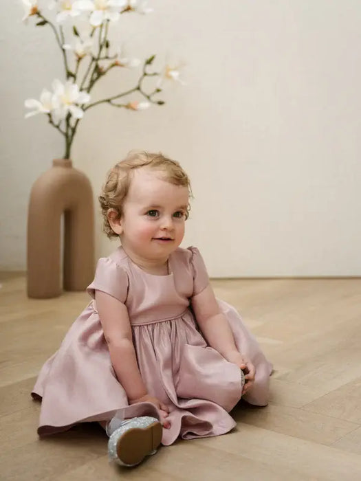 Teter Warm Harper pink baby party dress worn by a toddler sitting on a wooden floor indoors.